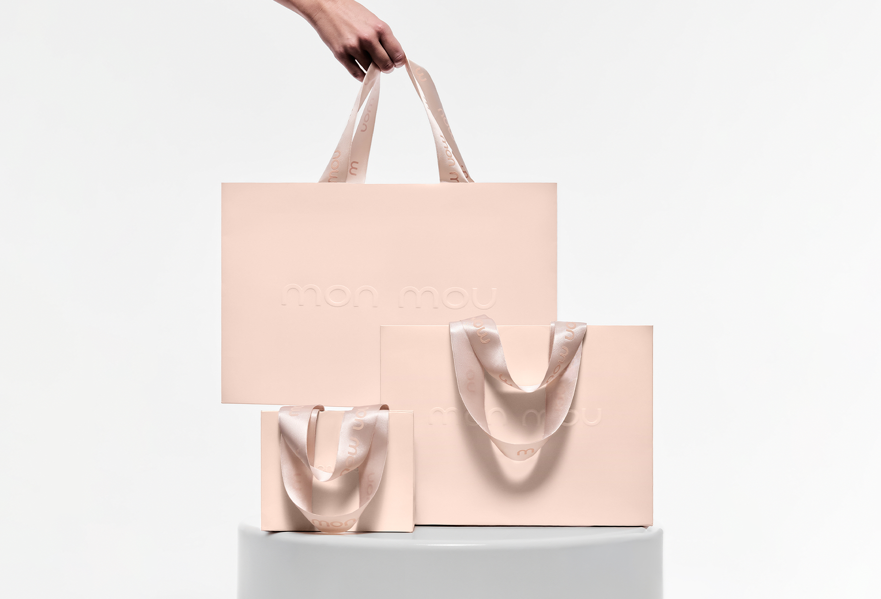 Stack of pastel pink gift bags with pale pink satin ribbons displayed on a white pedestal; embossed 'mon mou' on bags.