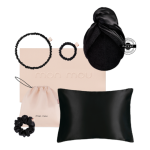 Flat lay of Mon Mou hair accessories set with two black scrunchies, a beige drawstring bag, a pale pink box, a black satin pillow, and a silk bonnet with a patent logo tag.