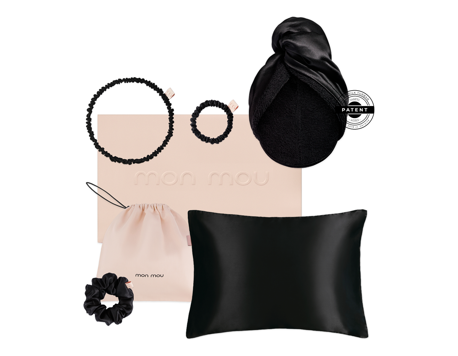 Flat lay of Mon Mou hair accessories set with two black scrunchies, a beige drawstring bag, a pale pink box, a black satin pillow, and a silk bonnet with a patent logo tag.