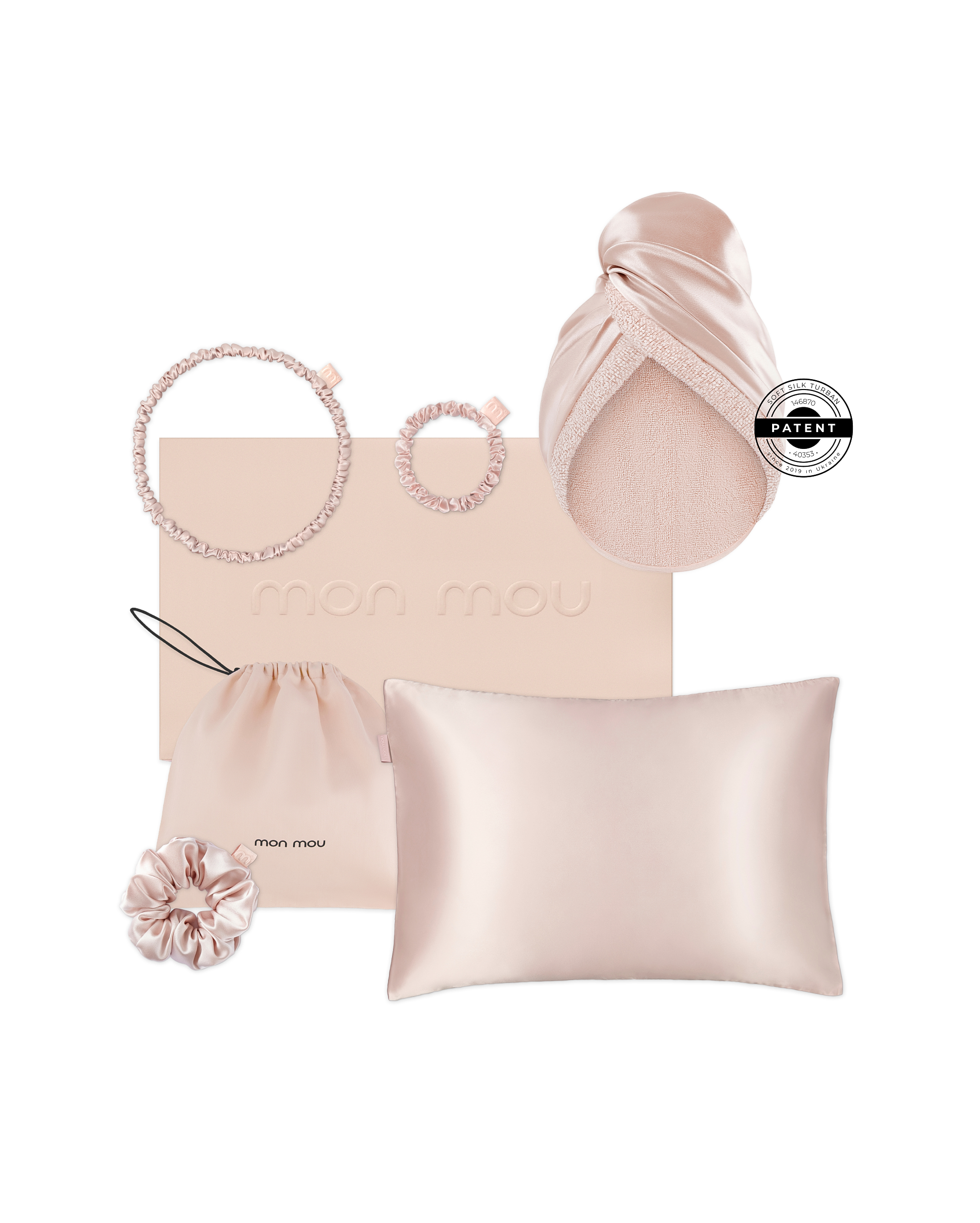 Soft pink beauty accessories arrangement: satin scrunchies, a silk head wrap draped over a curved surface, a blush pink padded pillow, and a pale pink box embossed with 'mon mou' on a white background.