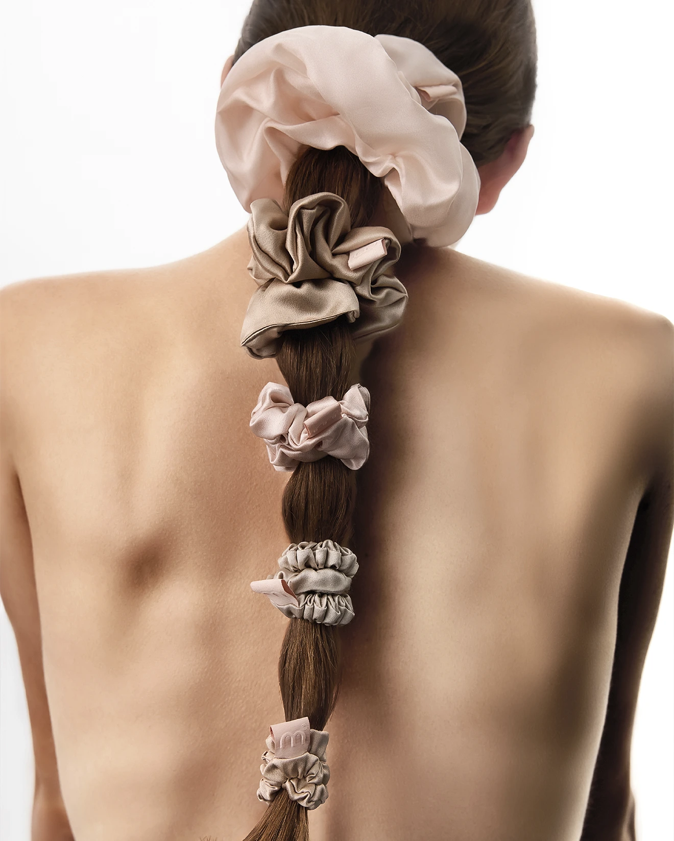 Back view of a woman with a long braided ponytail tied with soft pink satin scrunchies along the braid set against bare shoulders.