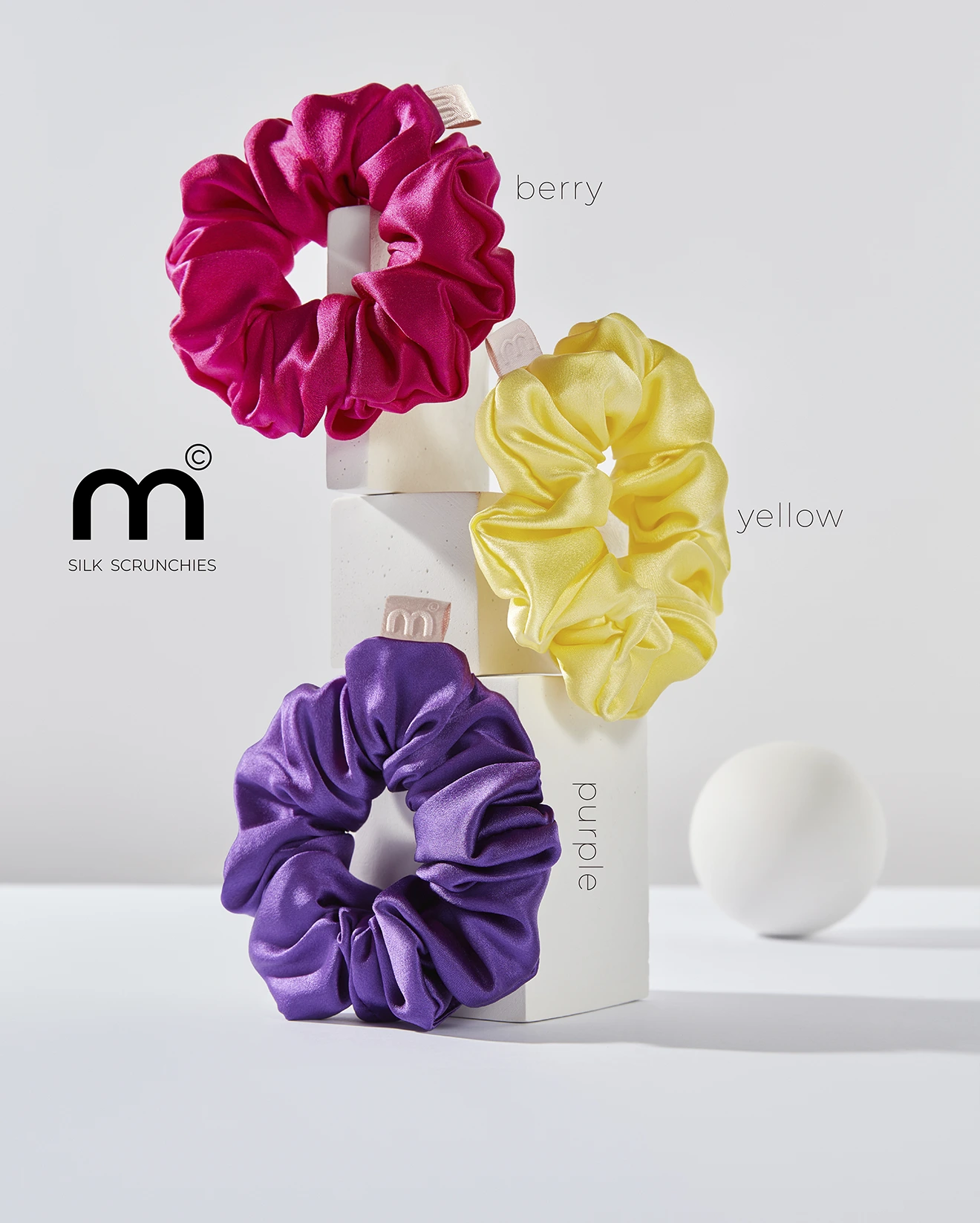 Product shot of colorful silk scrunchies (pink, yellow, purple) displayed on white blocks for a Silk Scrunchies brand image.