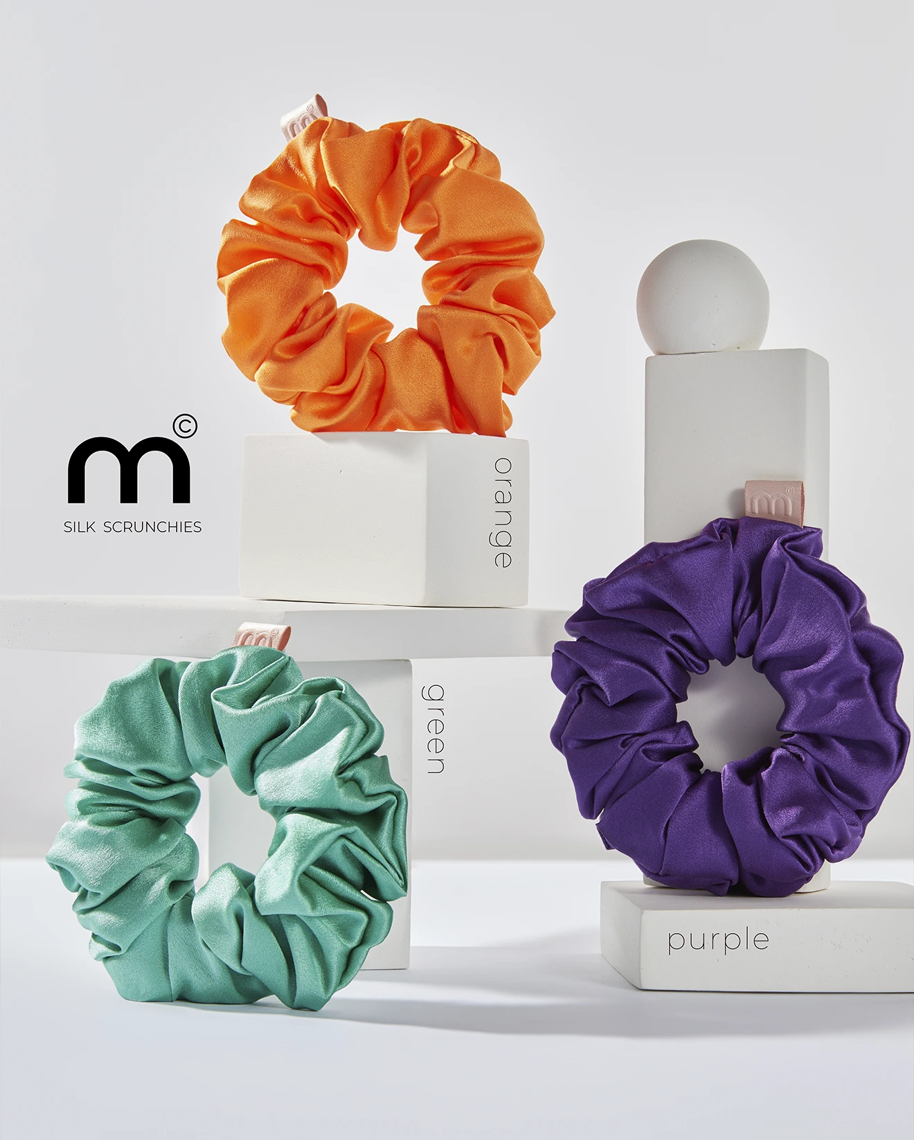 Colorful satin scrunchies in orange, green, and purple arranged on white display blocks with the 'm Silk Scrunchies' branding visible.