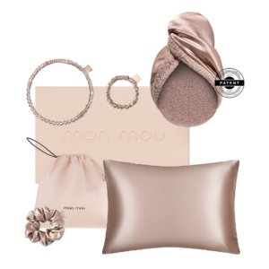 Flat lay of blush-pink accessories arranged on a pale pink box: two spiral hair ties, a satin scrunchie, a drawstring pouch, and a satin pillow; a round patent seal is visible on the right edge of a soft scarf.