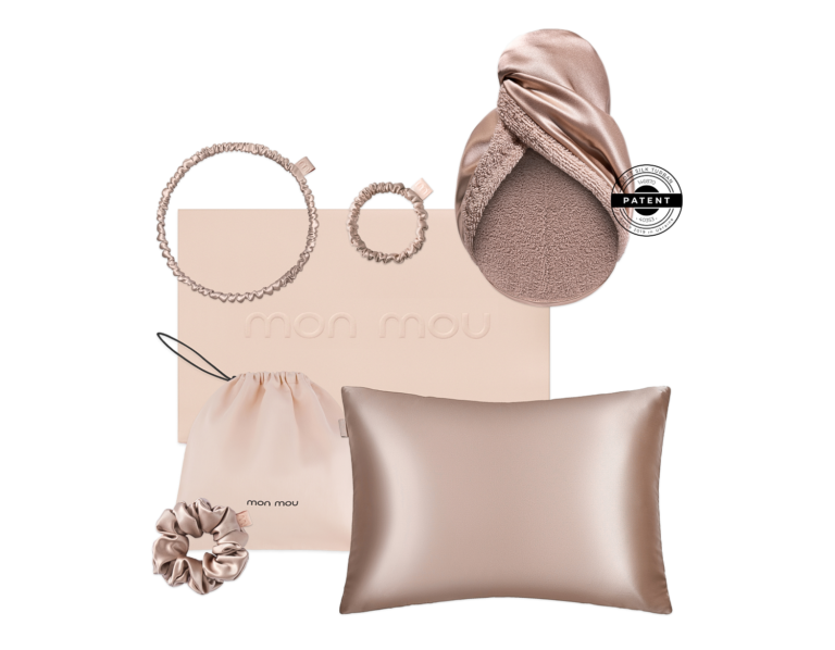 Flat lay of blush-pink accessories arranged on a pale pink box: two spiral hair ties, a satin scrunchie, a drawstring pouch, and a satin pillow; a round patent seal is visible on the right edge of a soft scarf.