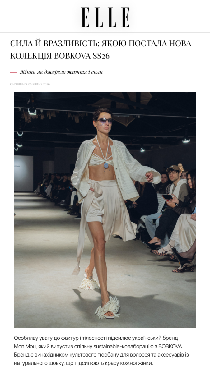 Model walks a runway in a beige two-piece outfit with a flowing robe, wearing sunglasses during a fashion show; audience seated along the catwalk.