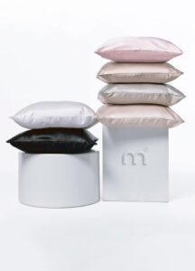 Stack of pastel satin pillows arranged on white display stands: pink, beige, gray, and cream, with a black pillow on a round pedestal.