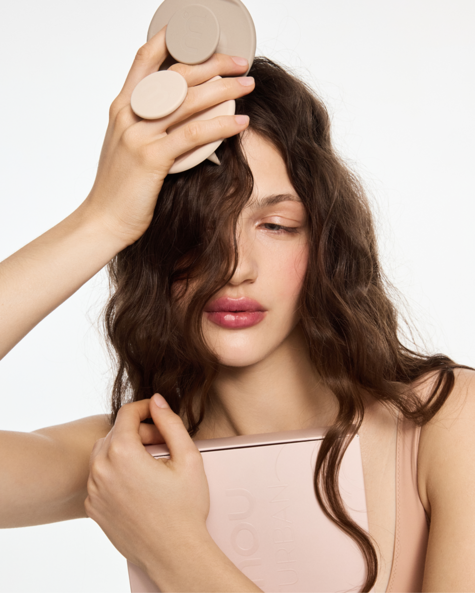 Woman with long brown hair holding a round beige beauty device against her scalp.