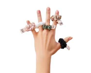 Hands showing several fabric scrunchies worn on fingers (index, middle, ring) against a white background, pink, white, green, beige colors.