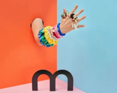 Close-up of a hand wearing colorful scrunchies and letter-bead bracelets, against a split orange and blue background.