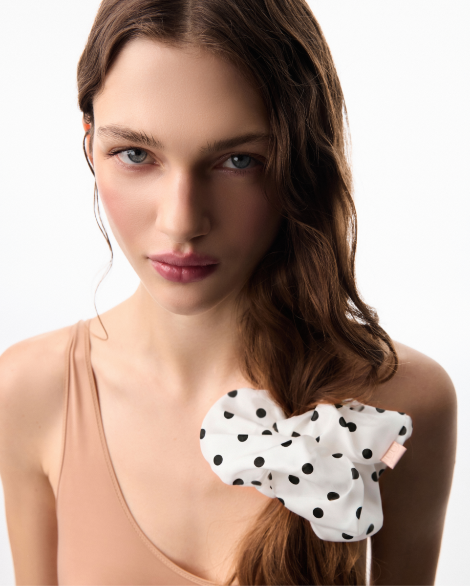 Close-up portrait of a woman with blue eyes, brown hair, wearing a peach top and a white polka-dot bow against a white background.