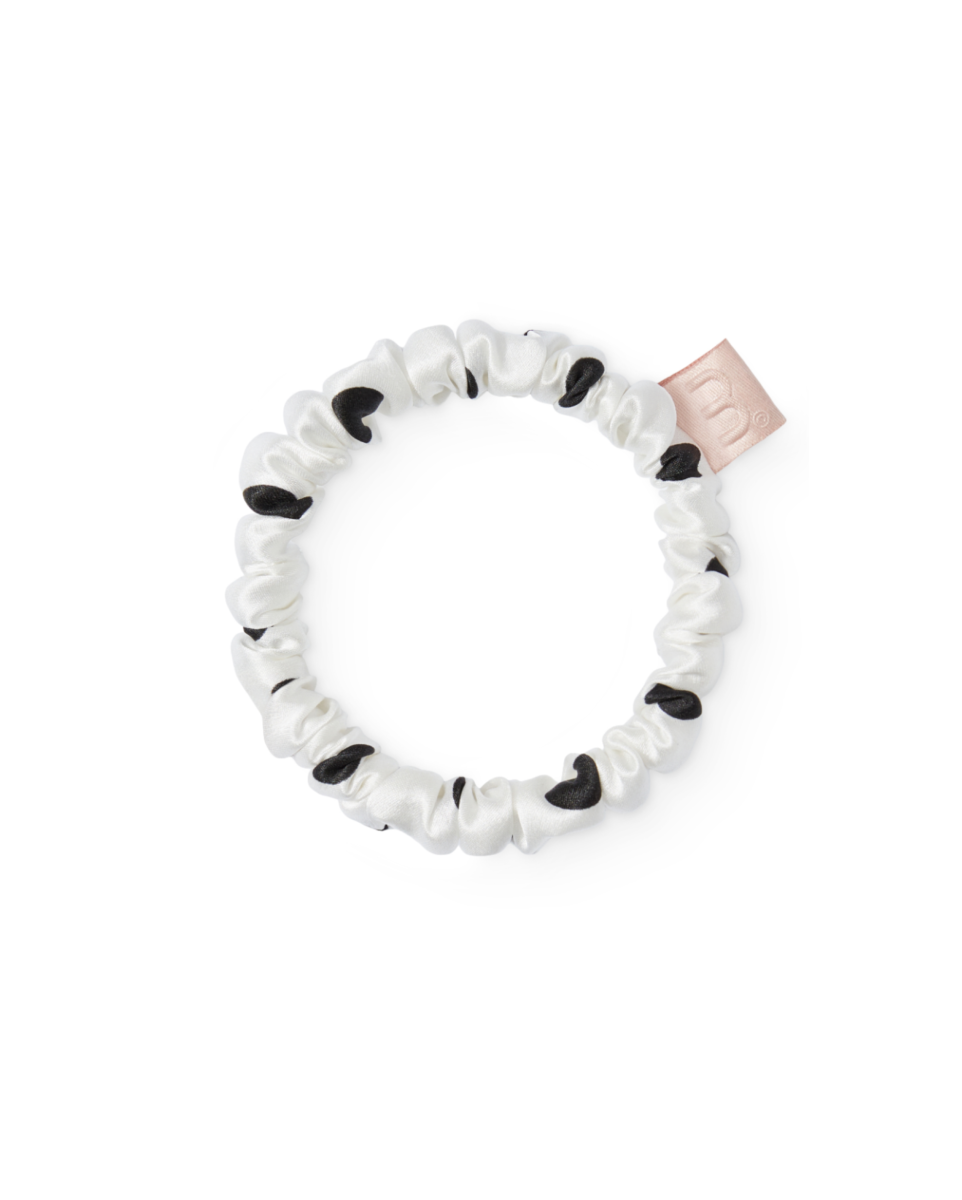 White satin scrunchie with black polka dots and a pale pink brand tag, shown in a close-up.