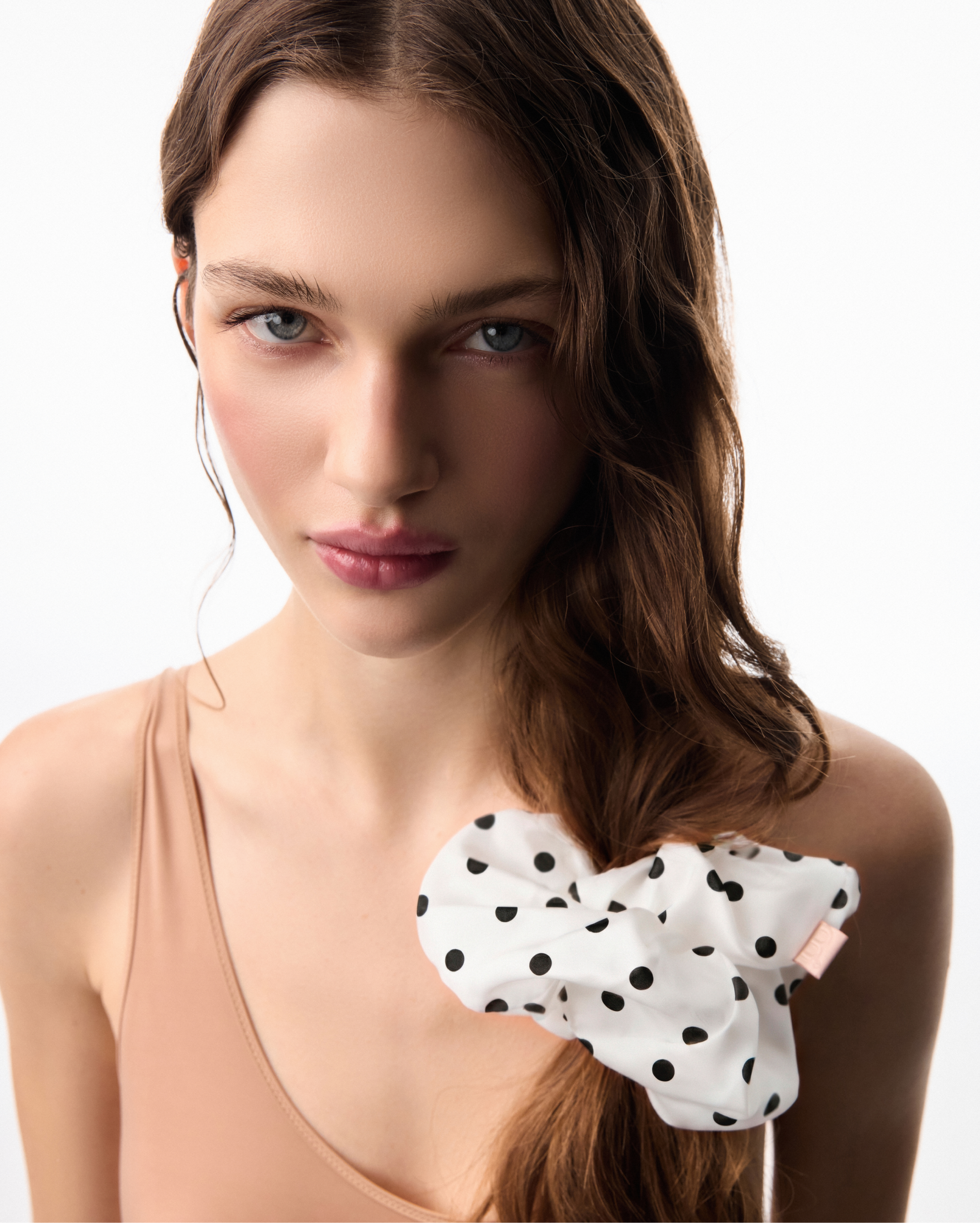 Close-up portrait of a woman with blue eyes, brown hair, wearing a peach top and a white polka-dot bow against a white background.