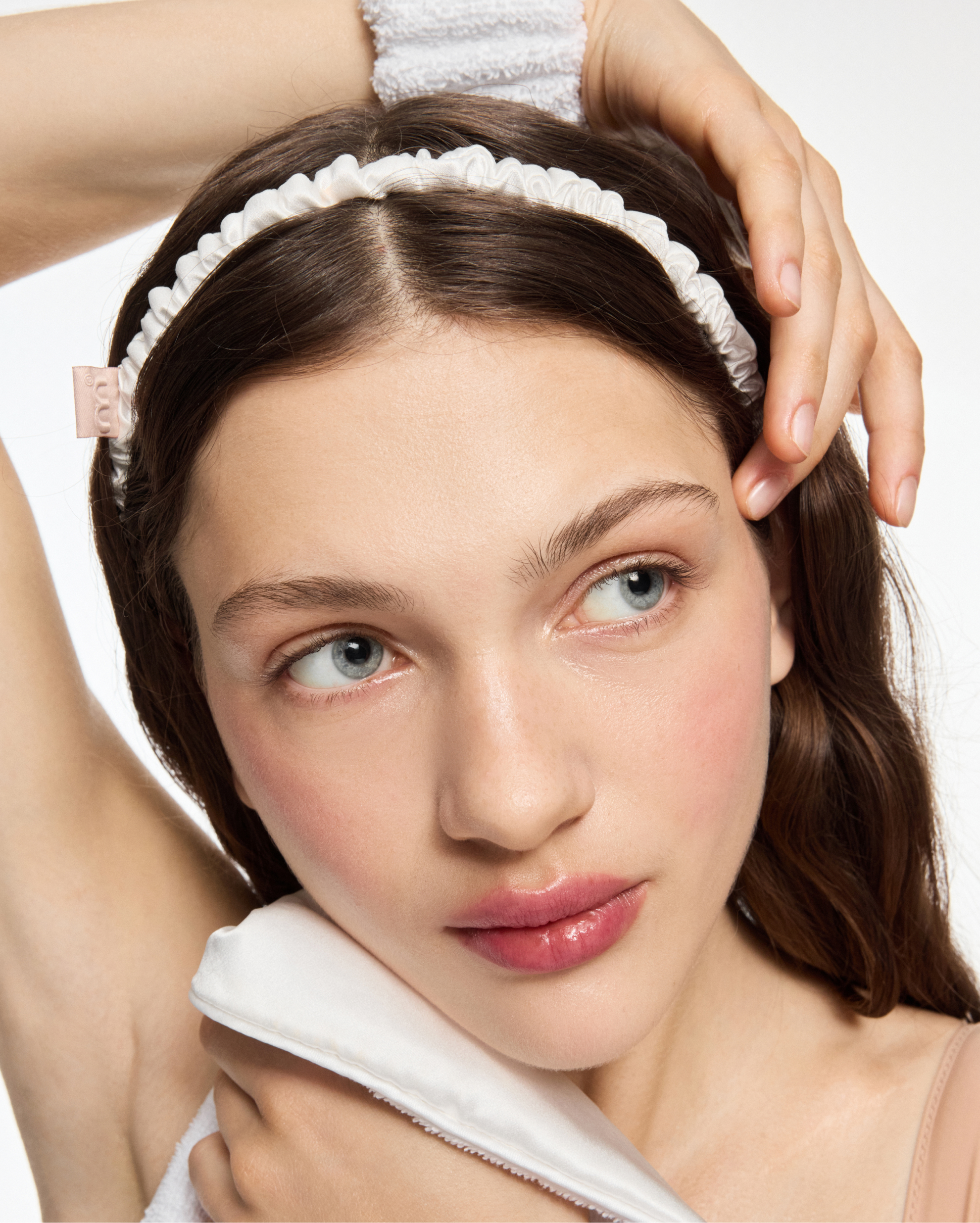 Close-up of a woman’s face with a white, ruffled headband and a soft scarf, as she rests her hand in her hair.