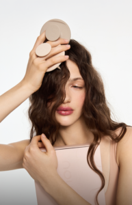 Woman holding a large beige circular beauty tool against her head, with a matching beige case nearby, in a pale pink outfit.