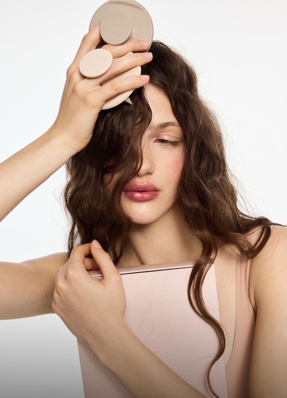 Portrait of a woman with wavy brown hair, holding beige circular makeup pads against her forehead and clutching a pink makeup palette near her chest.