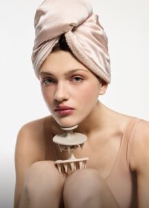 Woman with a soft pink towel wrapped in a turban, holding a beige facial massager against her chin in a studio portrait on a neutral background.
