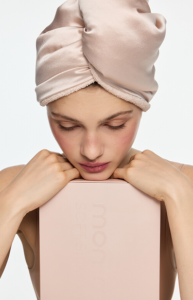 Woman with a pink satin head wrap holding a pale pink box close to her chest, looking down at it.