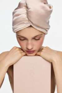 Close-up of a woman wearing a pale pink satin head wrap, resting her chin on a pale pink box with eyes closed.