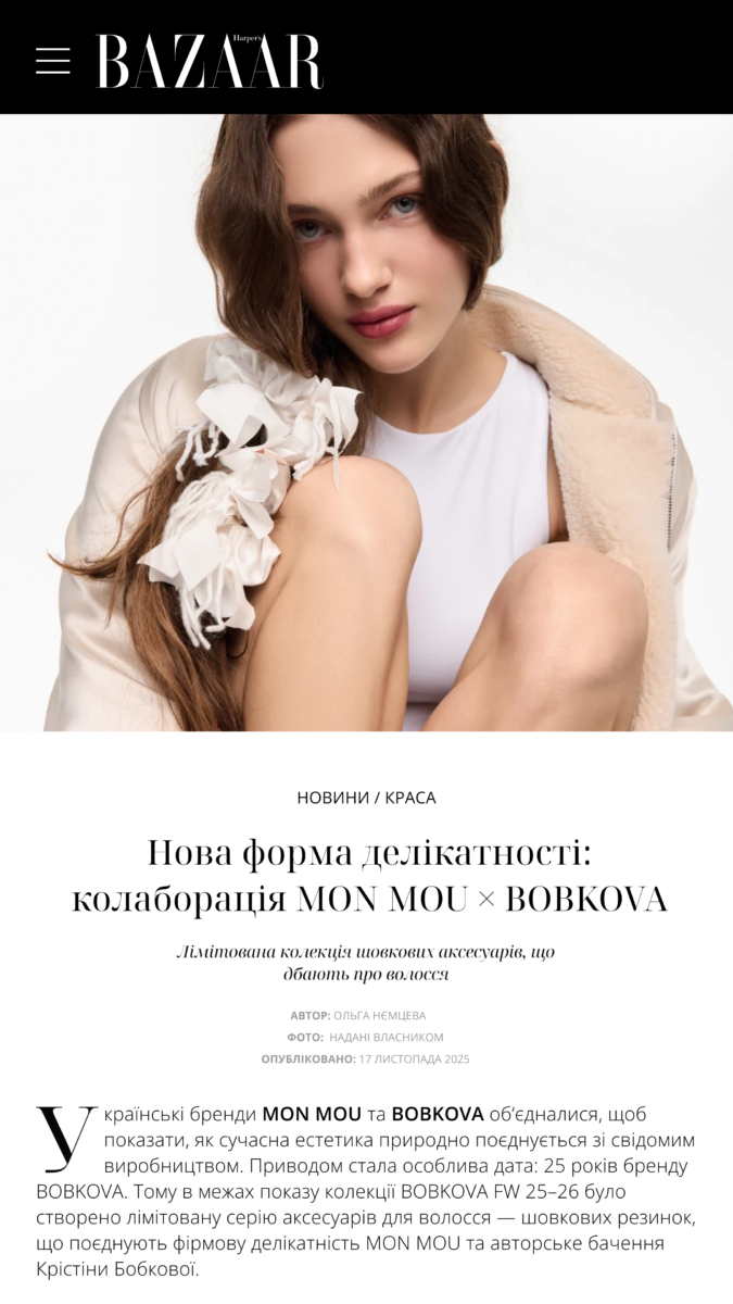 Portrait of a young woman with long brown hair, wearing a white top and beige shearling jacket, looking at the camera, with a white fabric flower accessory in her hair against a white background.