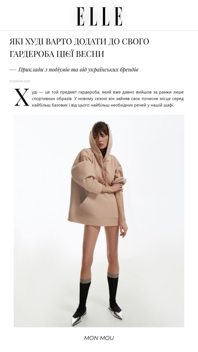 Model wearing a beige oversized hoodie dress with hood, posing against a white studio backdrop.