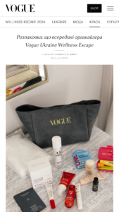 Flat lay of beauty products (lipsticks, serums, creams) scattered on a marble surface, with a dark Vogue tote bag in the background.