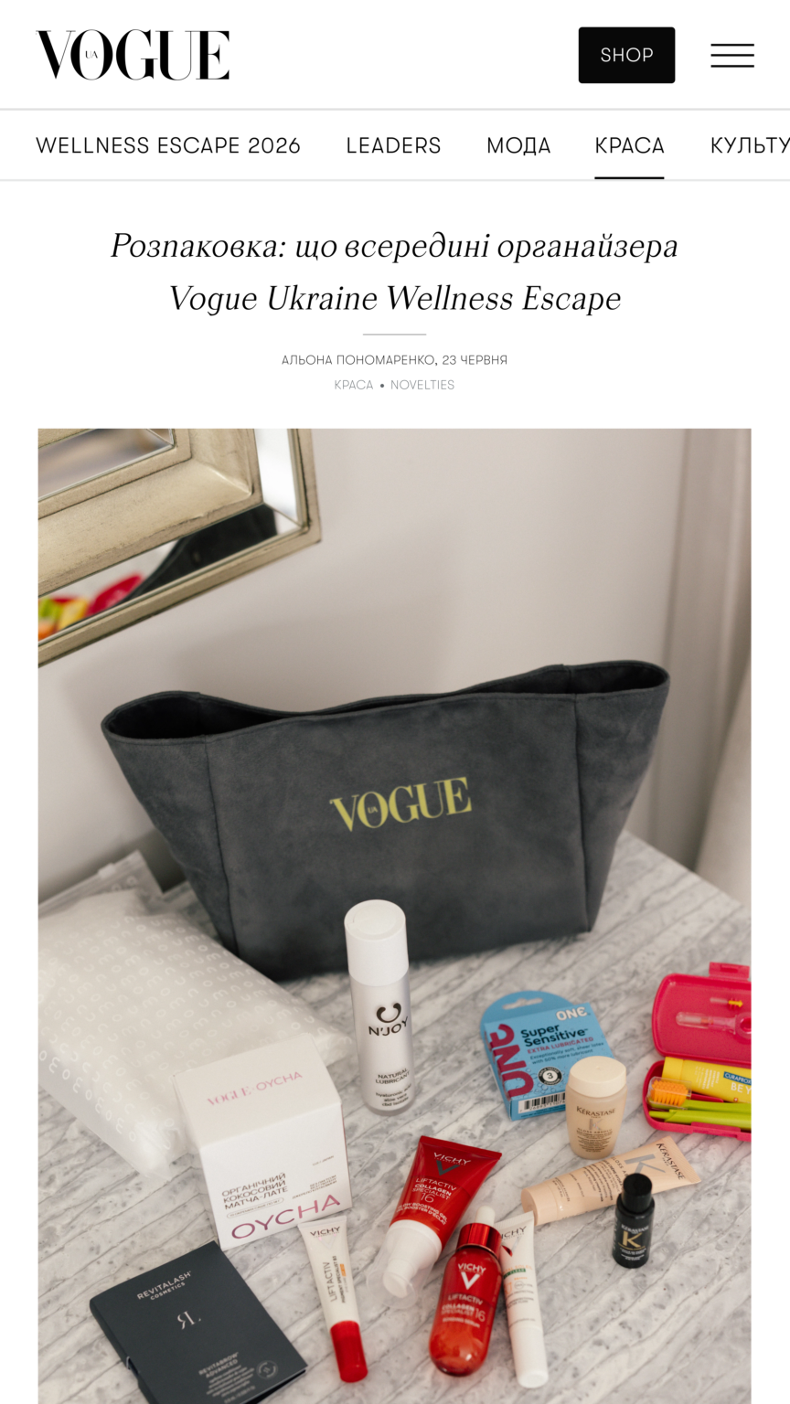Flat lay of beauty products (lipsticks, serums, creams) scattered on a marble surface, with a dark Vogue tote bag in the background.